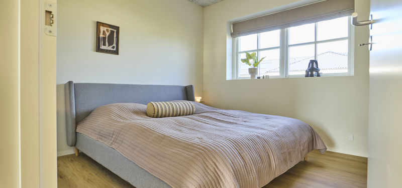 Lyst soveværelse fra eurodan-huse med grå polstret seng, beige sengetæppe med striber, rundt pyntepude, stort sprossevindue med rullegardin, plante i vindueskarm og enkel vægkunst over sengen – inspiration til nybyggeri og rolige husdrømme med skandinavisk indretning.