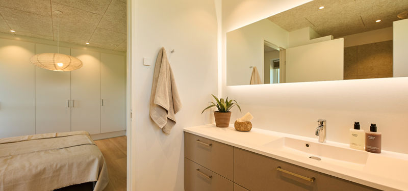 Badeværelse fra eurodan-huse med lyst vaskemøbel, stor spejlvæg, integreret vask, plante og kig ind til soveværelse – inspiration til elegant badeværelsesindretning, nybyggeri og husdrømme.