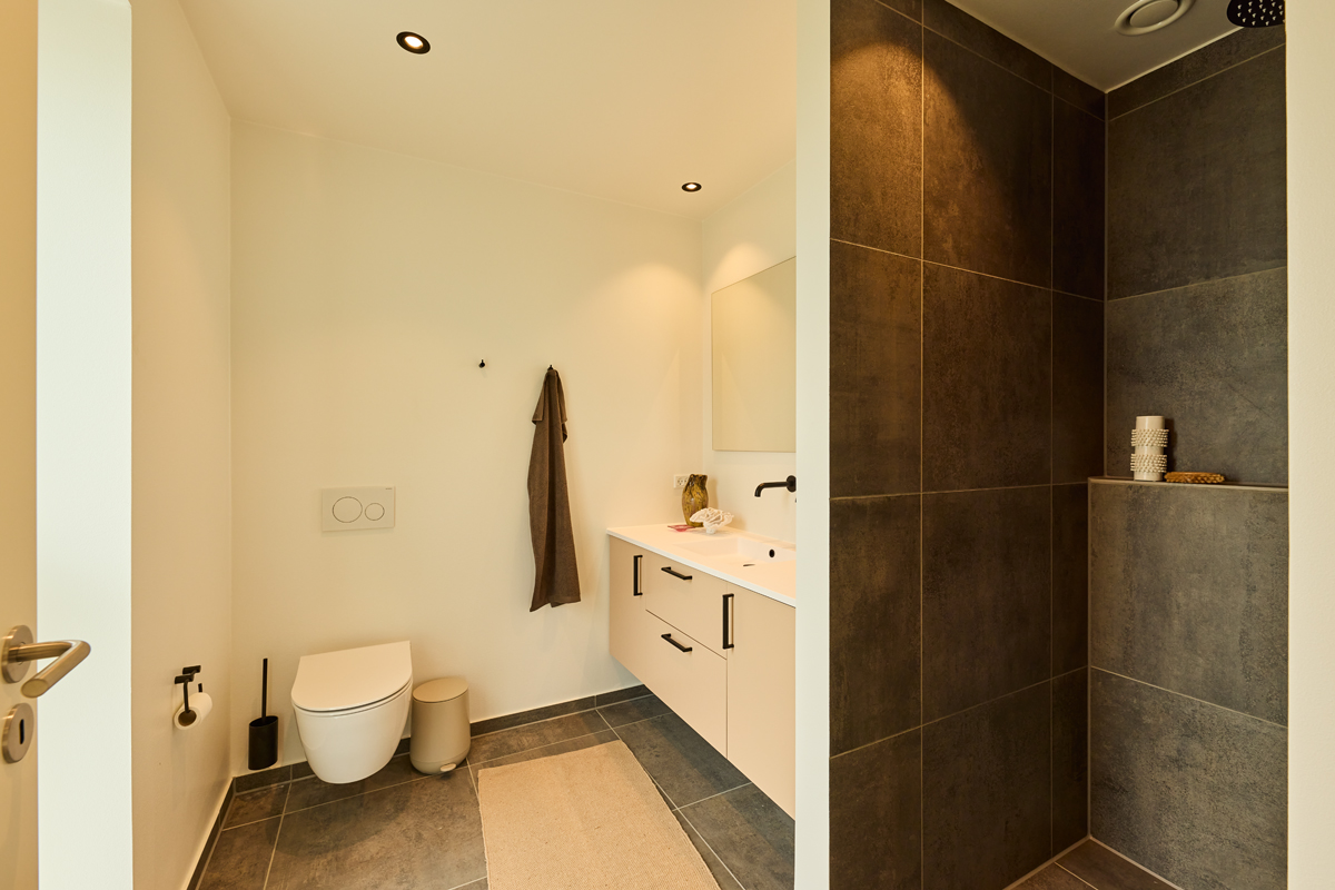 Badeværelse fra eurodan-huse med væghængt toilet, lyst badmøbel, sort armatur og bruseniche med mørke fliser og indbygget hylde – inspiration til nybyggeri.