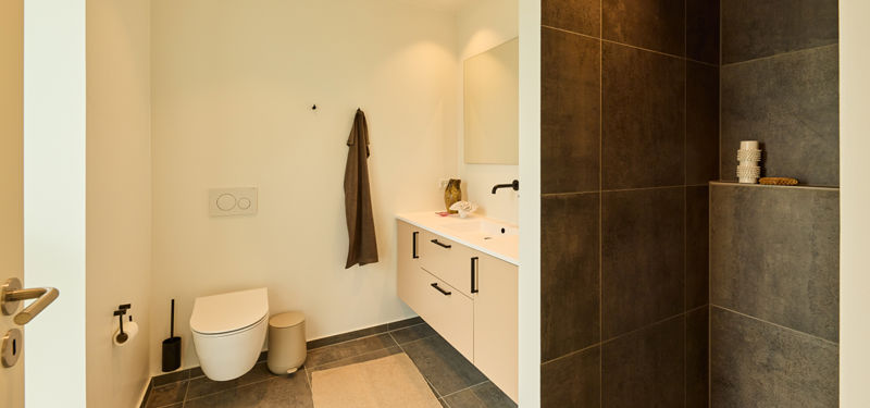 Badeværelse fra eurodan-huse med væghængt toilet, lyst badmøbel, sort armatur og bruseniche med mørke fliser og indbygget hylde – inspiration til nybyggeri.
