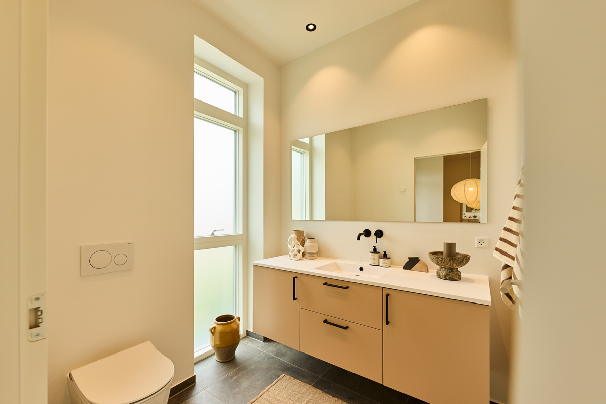 Badeværelse fra eurodan-huse med højt vindue, væghængt toilet, beige badmøbel, dobbelt vask, sort armatur og stort spejl – inspiration til stilrent nybyggeri.