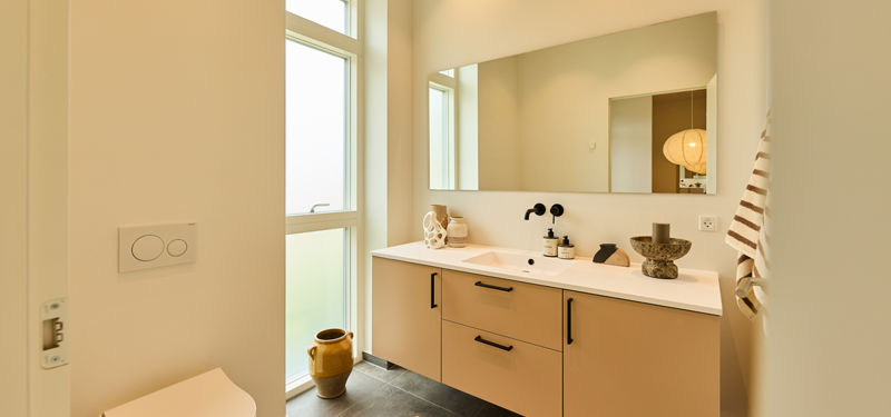 Badeværelse fra eurodan-huse med højt vindue, væghængt toilet, beige badmøbel, dobbelt vask, sort armatur og stort spejl – inspiration til stilrent nybyggeri.