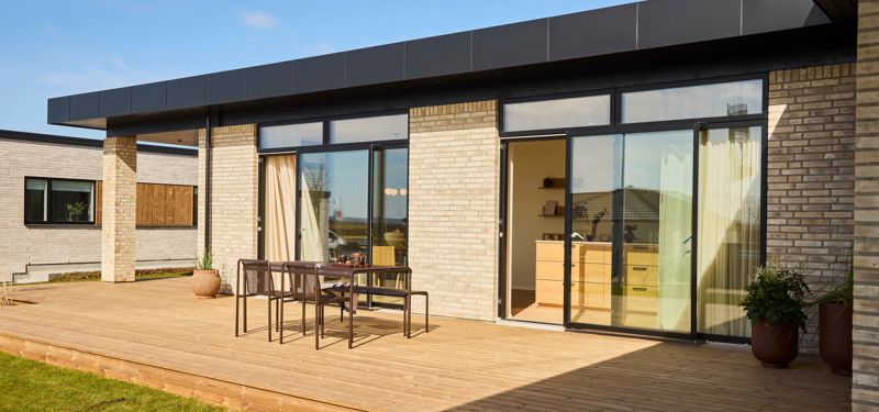 Træterrasse fra eurodan-huse foran lys murstensfacade med sorte vinduesrammer, åbne glasdøre, udendørs spisebord, potter ved facaden og direkte kig ind til køkken-alrum – inspiration til nybygget hus med terrasse, udeliv og gennemtænkt husbyggeri.