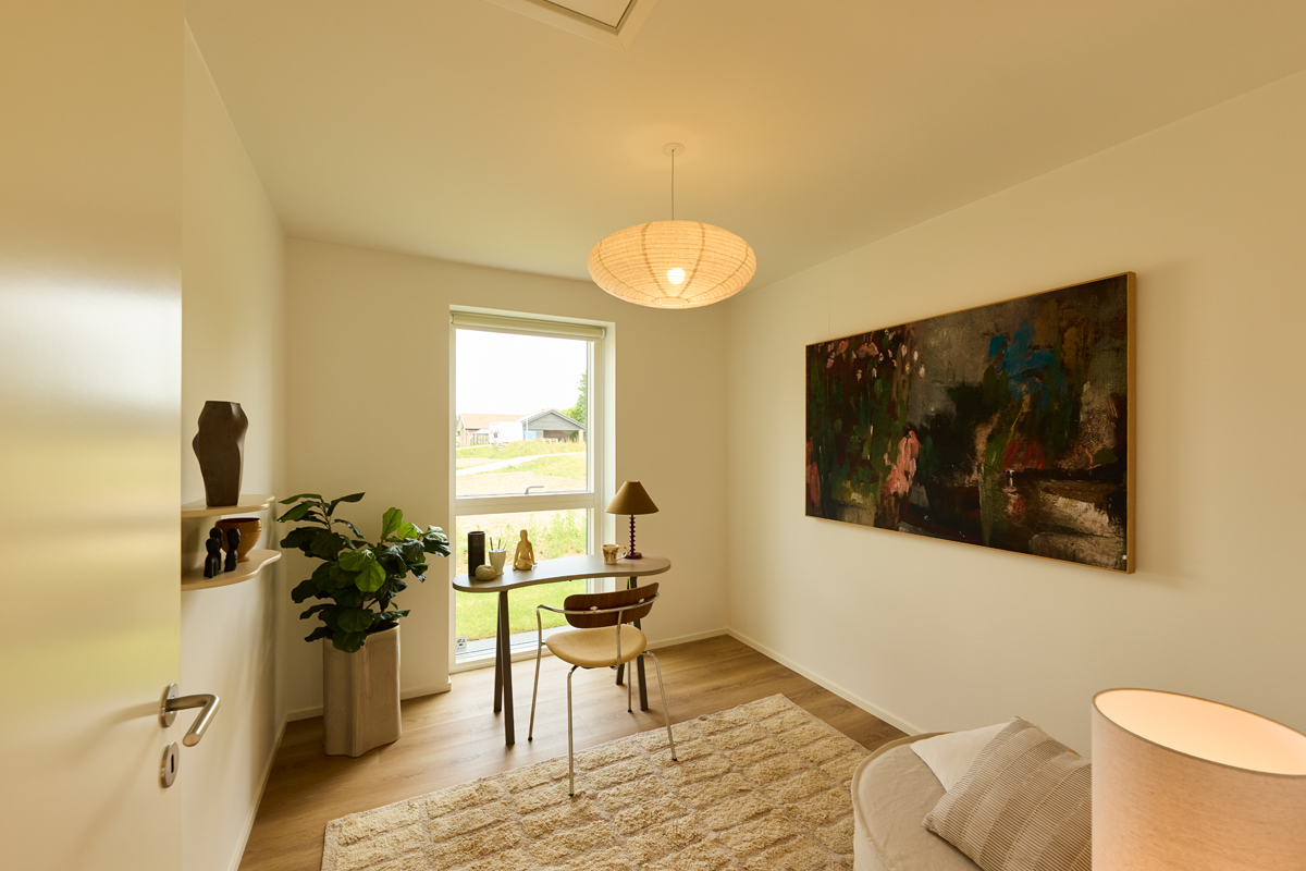 Kontor fra eurodan-huse med skrivebord ved vindue, loungestol, vægkunst, plante, gulvtæppe og rund pendellampe – inspiration til hjemmekontor i nybyggeri.