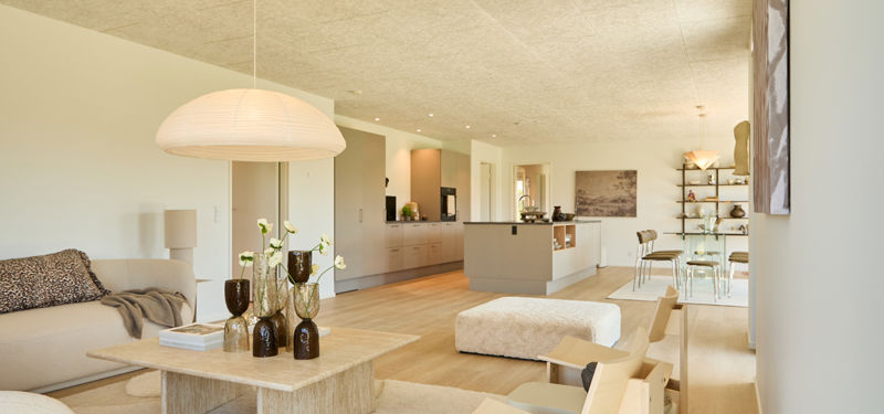 Kig fra stue mod køkken-alrum fra eurodan-huse med sofa, træsofabord, lys puf, køkkenø, beige køkkenfronter og spiseplads i baggrunden – inspiration til åben planløsning, nybyggeri og husdrømme.